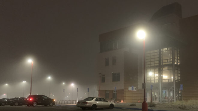Heavy fog over streetlights in a parking lot with a building in the background